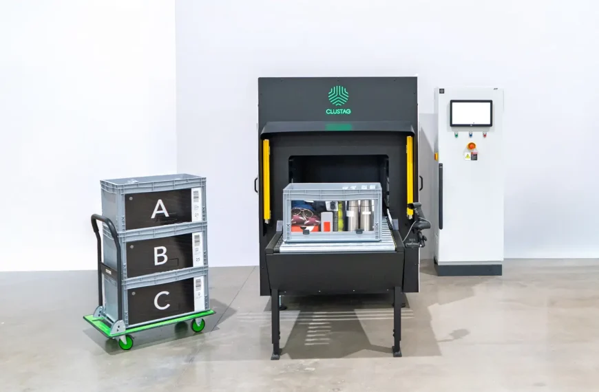 A gray storage bin containing assorted items—including sunglasses, a Ray‑Ban box, garments, and stainless‑steel bottles—sits on the conveyor inside a Clustag RFID Tunnel S24. To the left, three stacked crates labeled A, B, and C are visible
